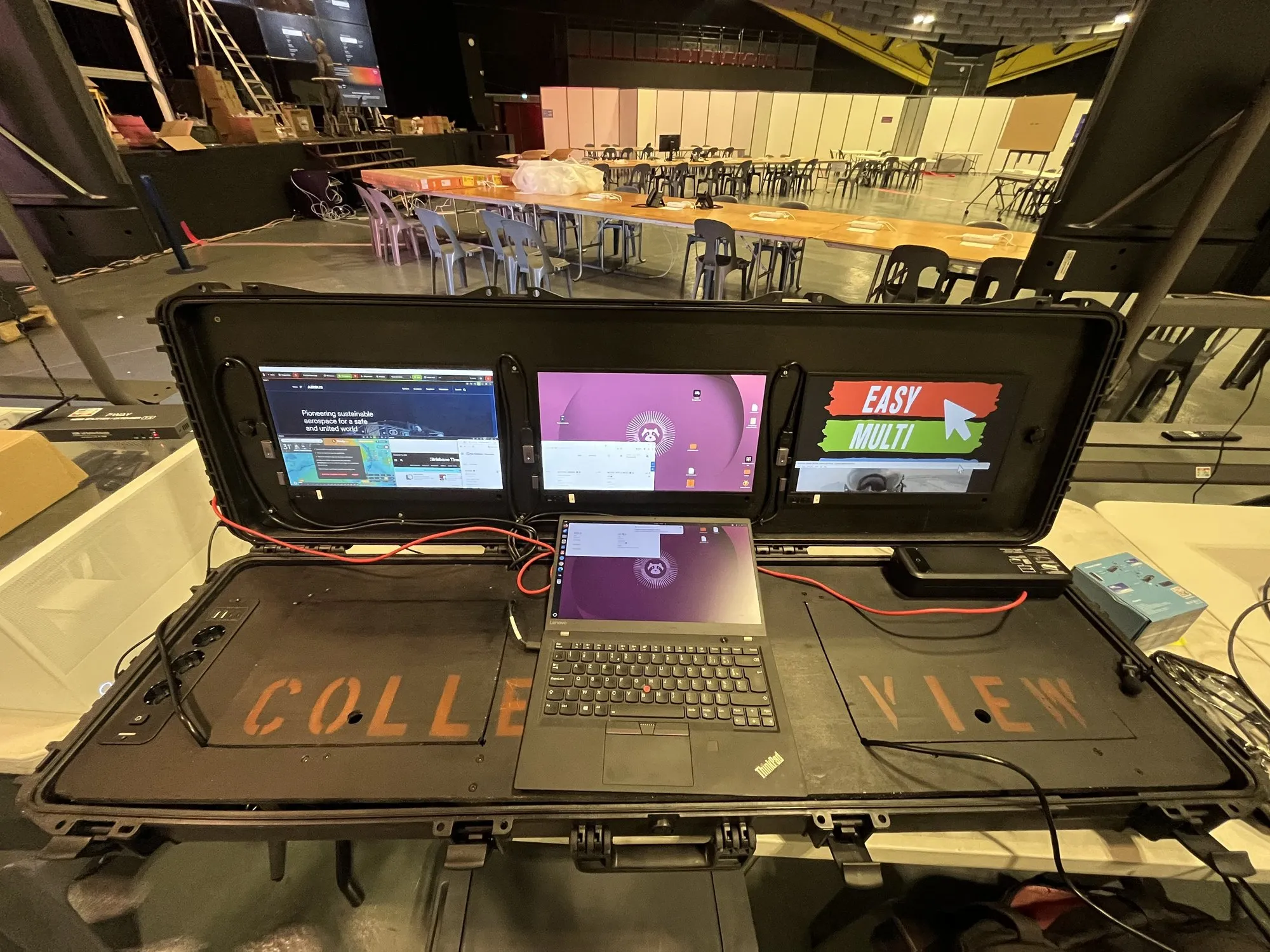 Maleta de mando EMD abierta en la sala de control de Lourdes 2026, el logotipo de Easy Multi Display en la pantalla, las mesas de los servicios al fondo