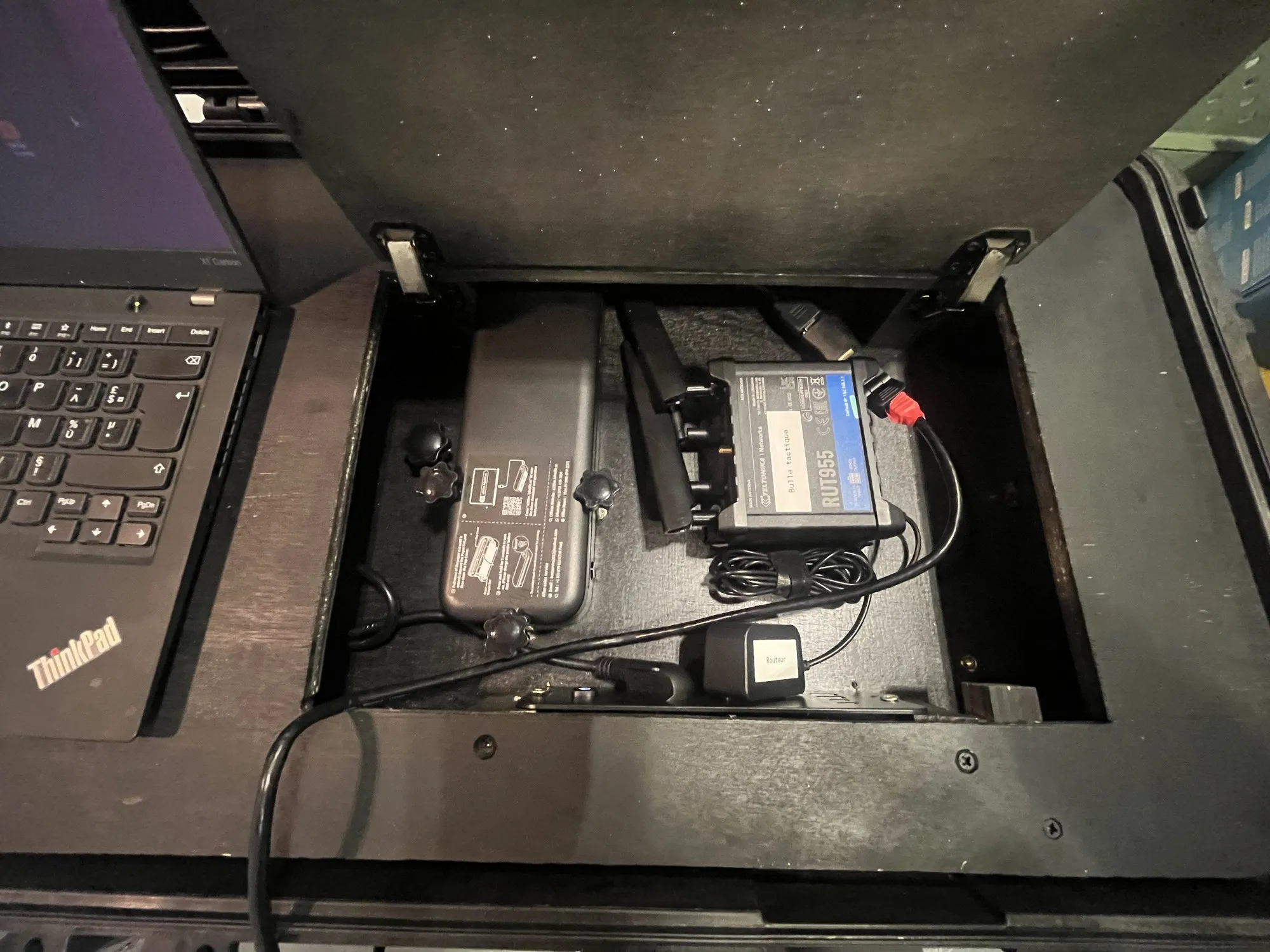 Trampilla técnica abierta bajo el plano de trabajo: router Teltonika, estación de acoplamiento y cables, el portátil a la izquierda