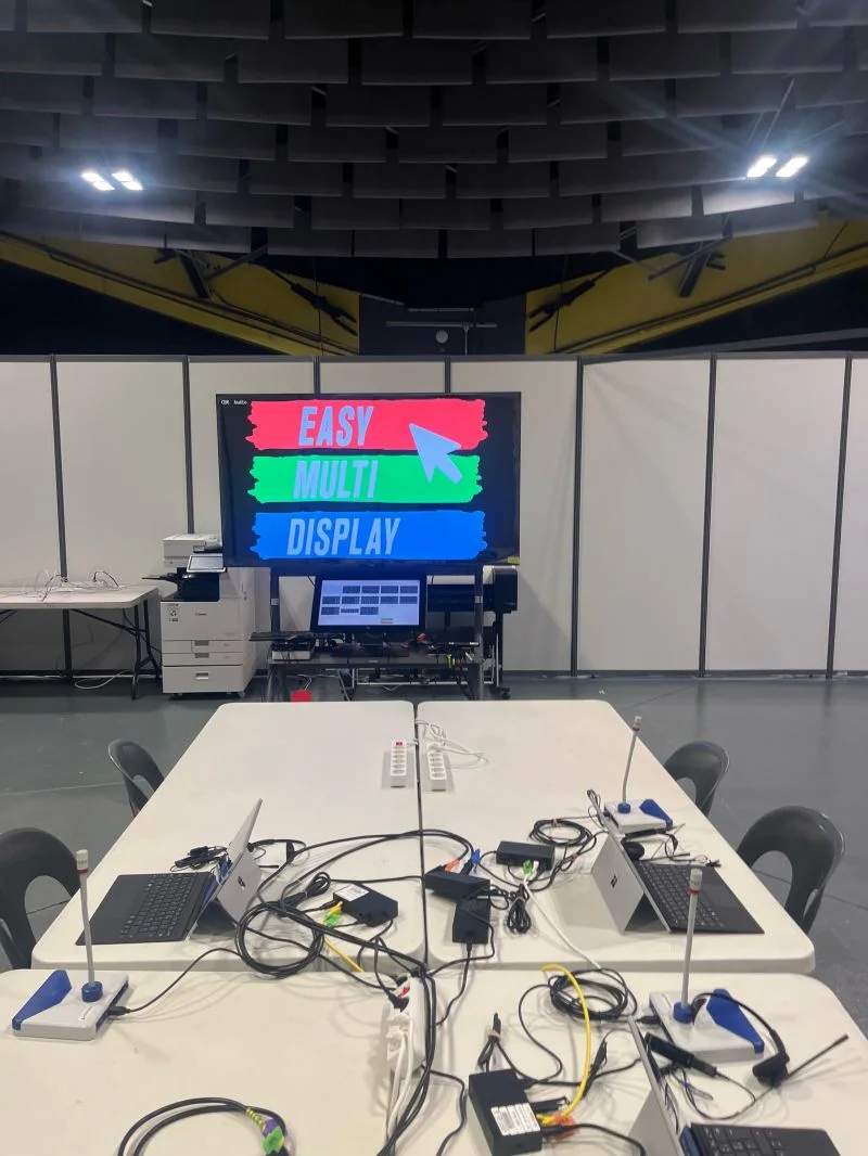 Vista di un'isola della sala: computer portatili e microfoni sui tavoli, schermata di benvenuto Easy Multi Display sullo sfondo